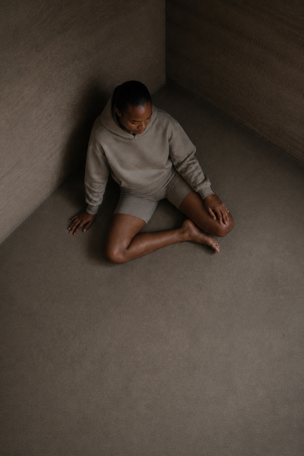 A barefoot woman in taupe clothing demonstrating a relaxed floor-sitting position at the corner of a rammed-earth room